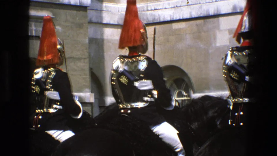 1969 England 8mm frame showing mounted ceremonial guards with red plumes and dark uniforms, partially framed by dark vertical obstructions.