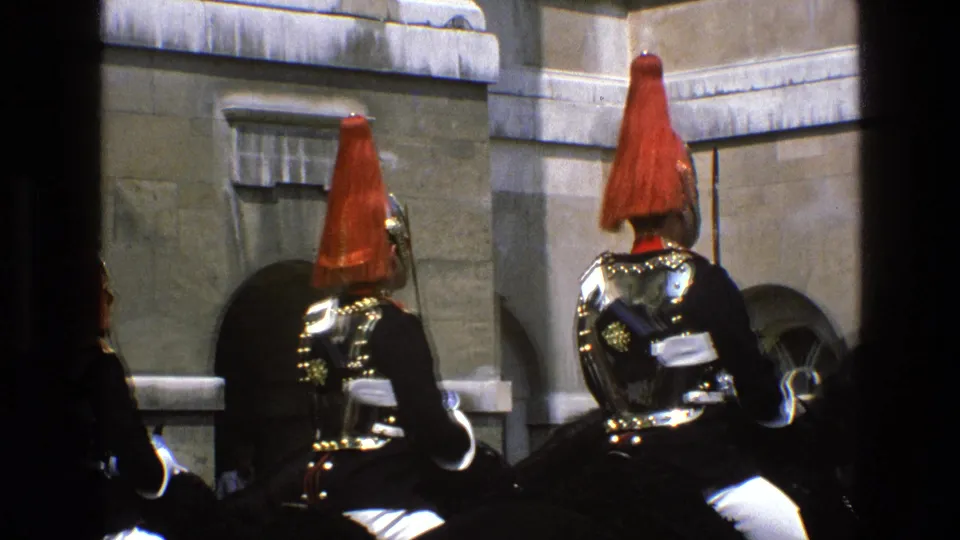1969 England home-movie frame of red-plumed mounted guards on black horses, with stone architecture and a window behind them.