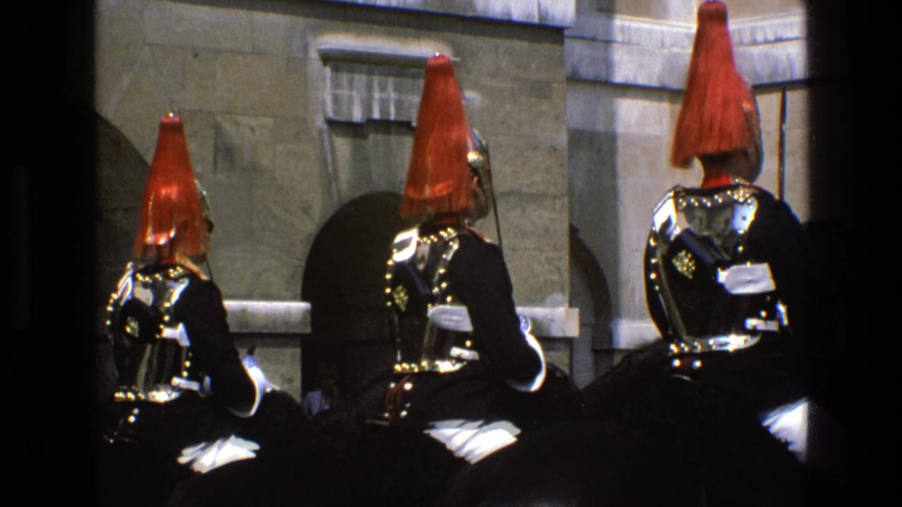 Mounted ceremonial soldiers in red plumed helmets pass a stone building with arched openings.