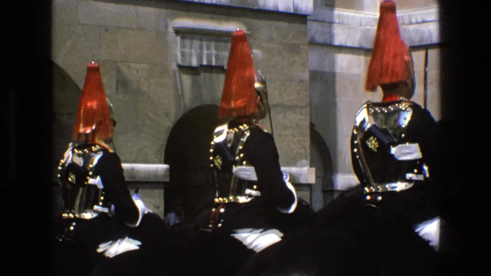 In 1969 England, mounted ceremonial soldiers in red plumed helmets and polished armor are seen from behind near stone arches.