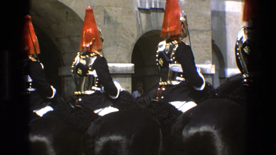 Mounted ceremonial soldiers in England wear red plumes, dark uniforms, and reflective armor while positioned near a stone facade.