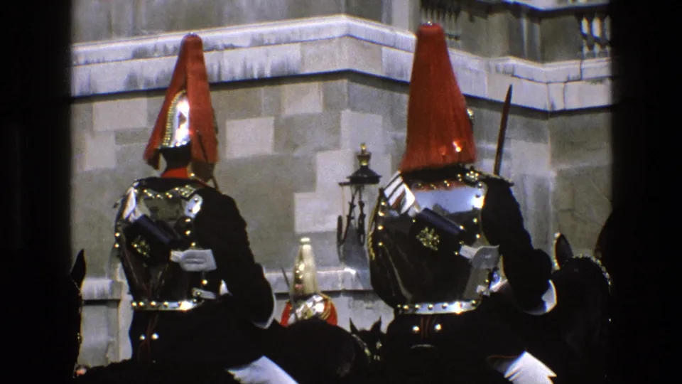 Two mounted ceremonial soldiers in red plumes and polished armor are framed against stone architecture in 1969 England footage.