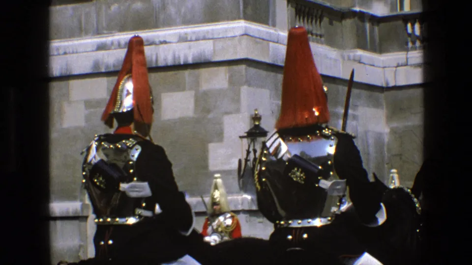 Two mounted guards in ceremonial dress are seen from behind in England, with red plumes, reflective armor and horses partly visible.