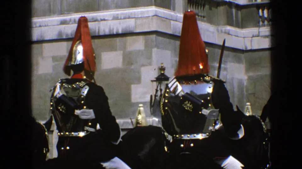 Visitor-shot 8mm frame of mounted ceremonial guards in England, with polished armor, red plumed helmets and a stone official-looking facade.