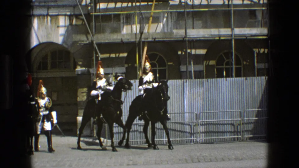 Ceremonial mounted guards in polished helmets and white gloves pause near metal barriers outside a scaffolded stone building.