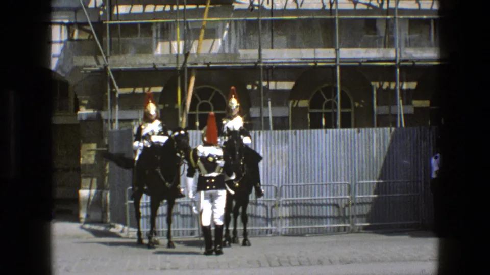1969 England home-movie frame showing a soldier between two mounted guards, with barriers and scaffolding behind them.