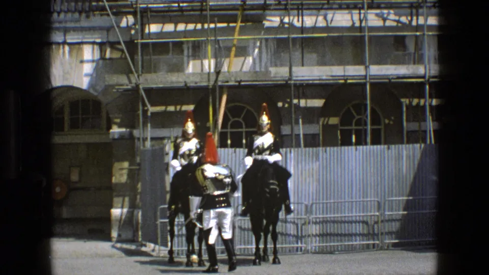 1969 England frame of mounted ceremonial guards and a soldier in dress uniform near a scaffolded building and crowd-control barriers.