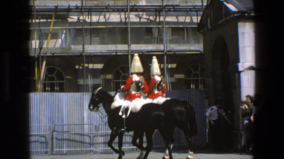 1969 England 8mm frame with two ceremonial mounted guards moving past barriers, spectators, scaffolding, and stone architecture.