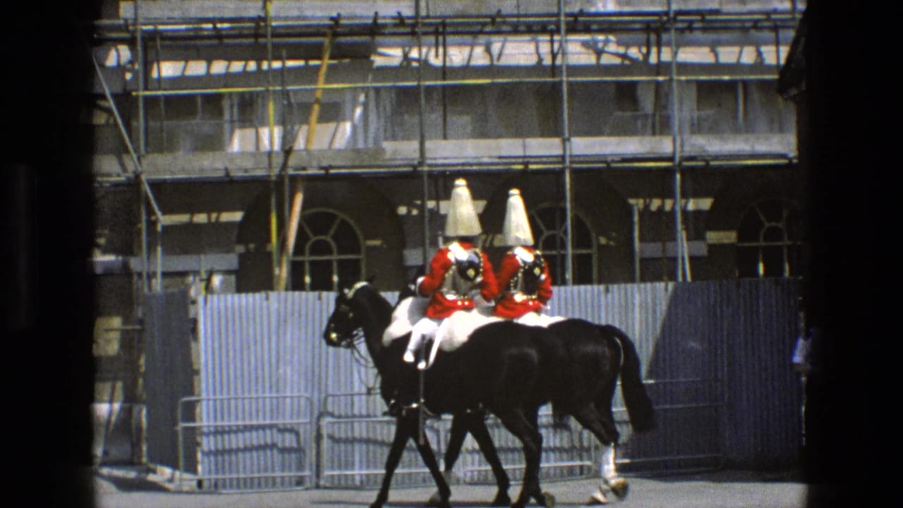 Two mounted guards in red coats ride black horses past a stone building with scaffolding.