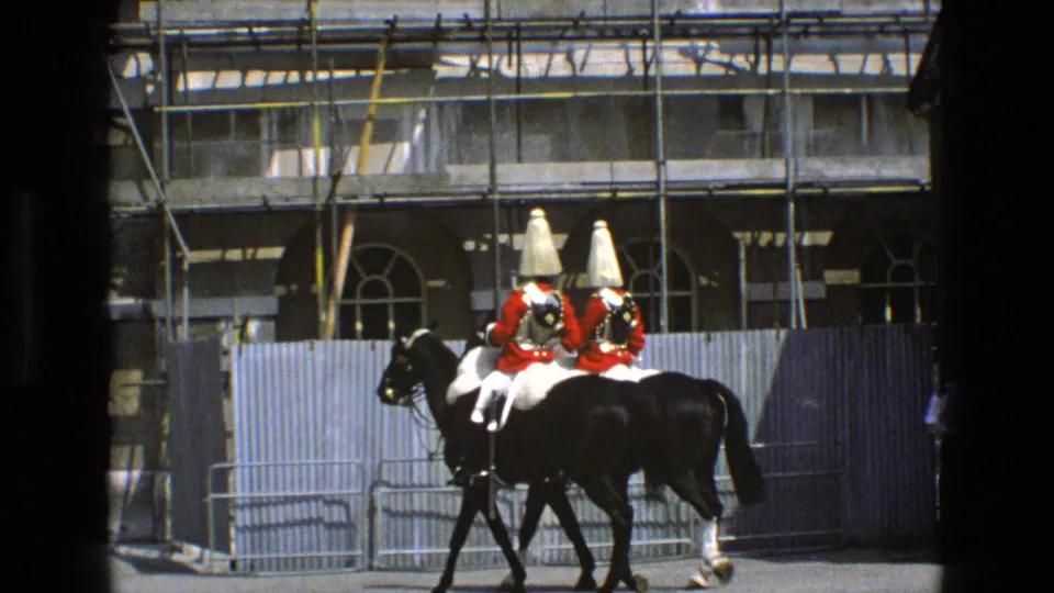In England archival 8mm footage from 1969, two ceremonial mounted guards in red coats ride black horses beside barriers and scaffolding.