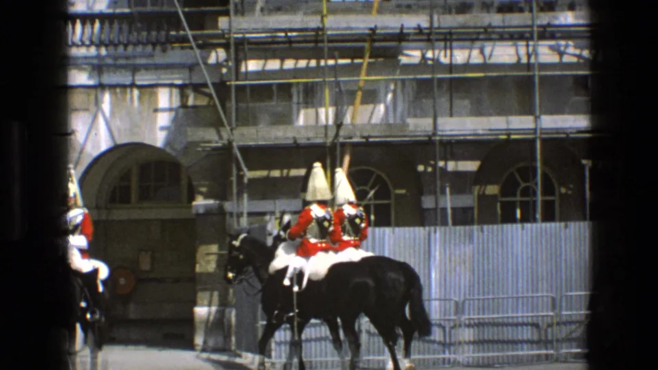 Ceremonial riders in red coats and tall gold-topped helmets pass arched stone doorways in England, filmed on 8mm in 1969.