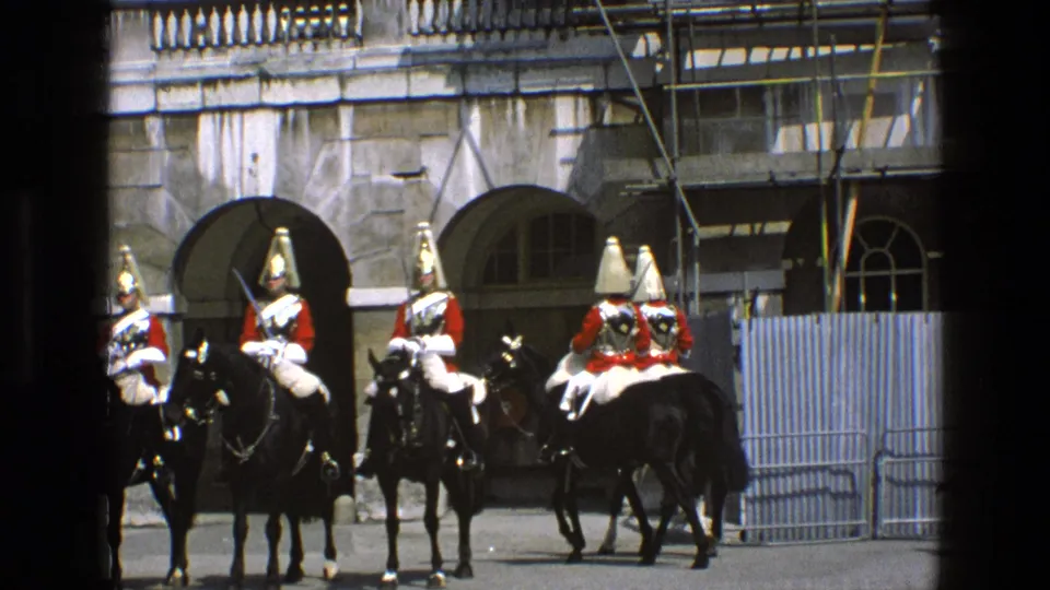 A group of mounted ceremonial guards in red coats and pale breeches is seen near stone arches and barriers in 1969 England footage.