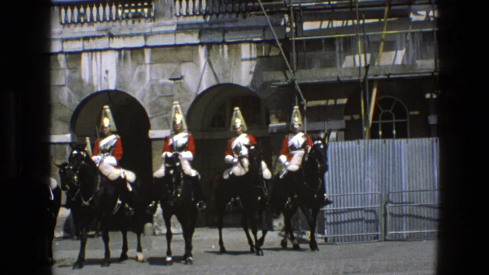 In England in 1969, uniformed cavalry in red tunics and polished helmets stand mounted on black horses near barriers and a scaffolded stone facade.