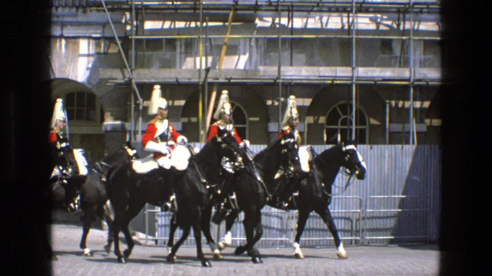 In England in 1969, ceremonial cavalry on black horses move laterally across a paved area near crowd barriers and a scaffolded stone building.
