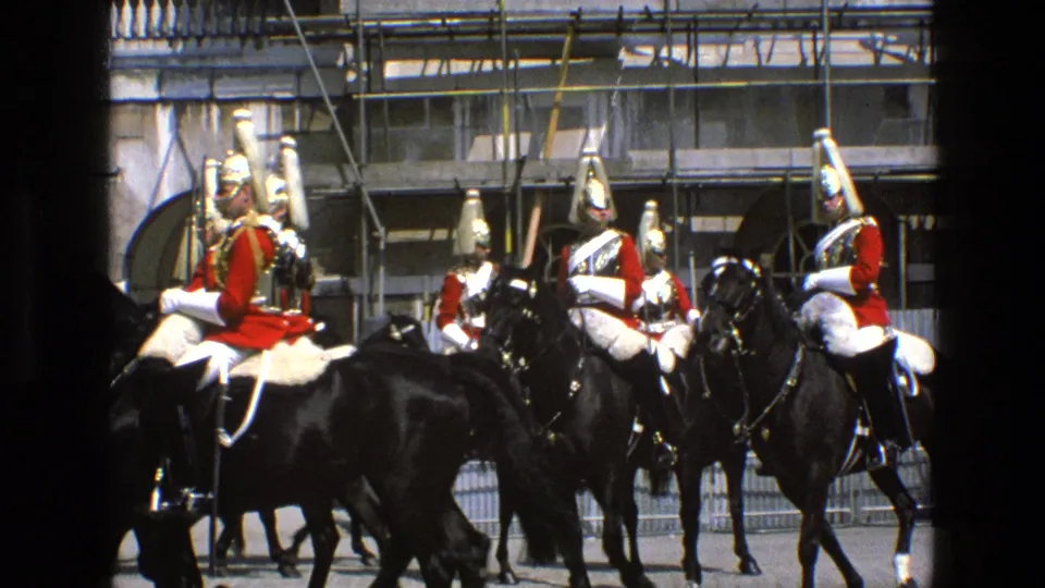 In 1969 England footage, mounted ceremonial guards in red tunics and plumed helmets ride black horses past stone arches.