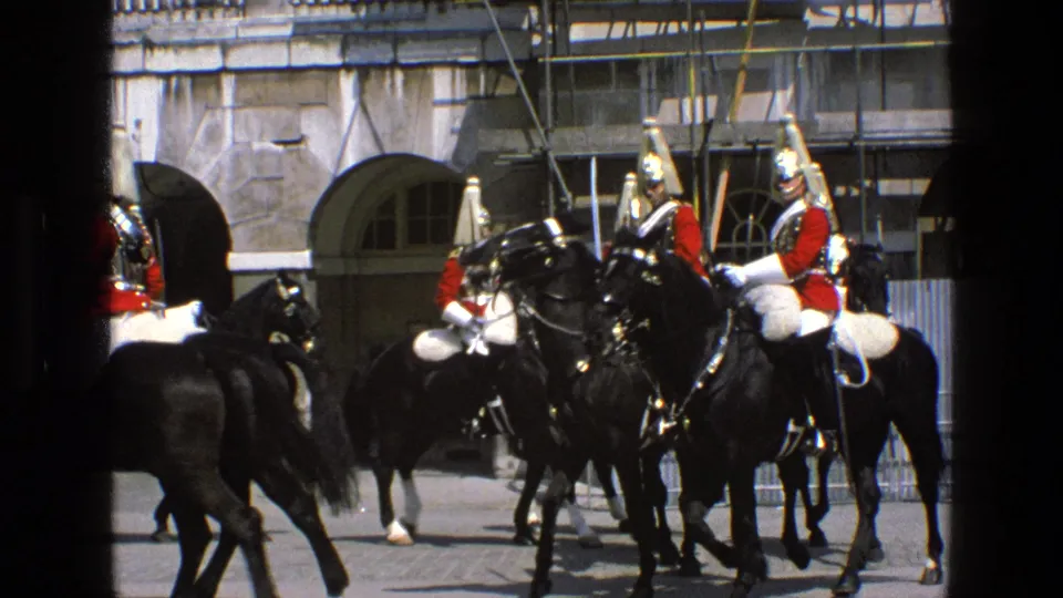 A handheld 8mm frame shows mounted ceremonial guards and black horses grouped near railings and stone arches in England, 1969.