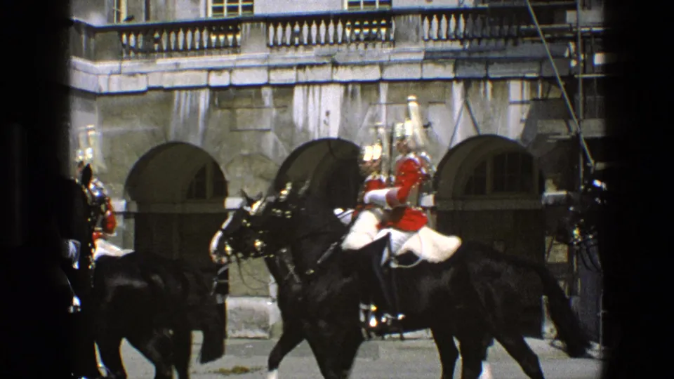 In 1969 England footage, mounted guards in red and dark uniforms pass a stone building with arched openings, partly framed by dark foreground posts.