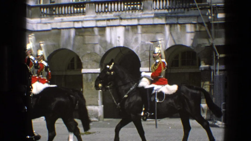 In 1969 England footage, a red-uniformed mounted guard on a black horse moves past a grand stone wall with arched openings.
