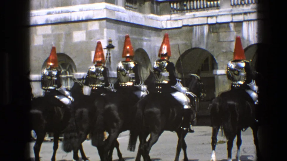 In 1969 England footage, a formation of mounted guards in dark uniforms and tall red helmets is seen from behind near arched stone doorways.