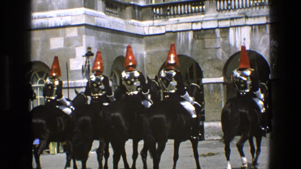 1969 England home-movie frame showing mounted ceremonial guards with red plumed helmets and polished armor outside stone arches.