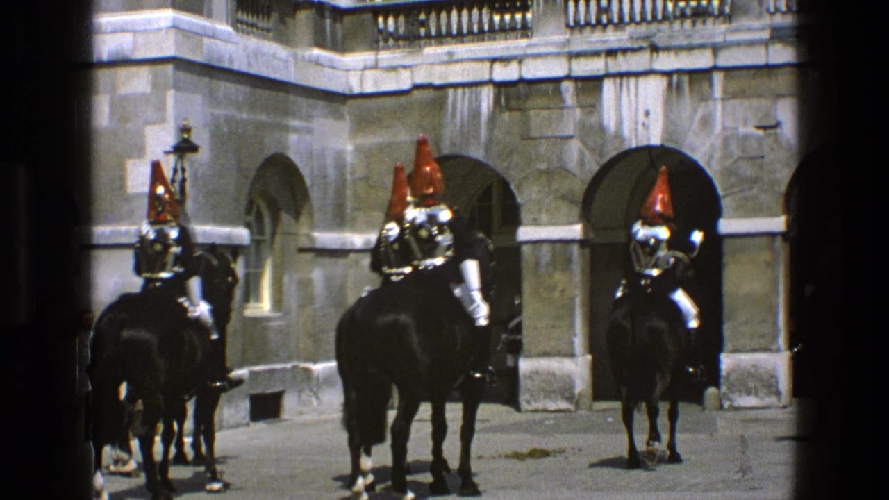 1969 England home-movie frame showing mounted ceremonial guards with red plumes in a palace-like stone courtyard.
