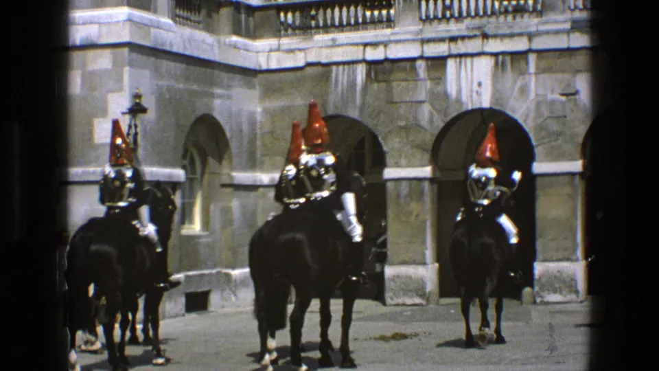 1969 England home-movie frame showing mounted ceremonial guards with red plumes in a palace-like stone courtyard.