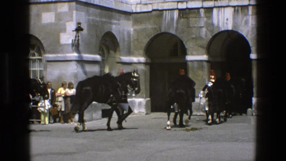 A black horse moves across the courtyard in 1969 England as uniformed guards and other horses stand near stone archways.