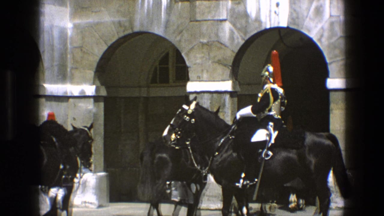 1969 England home-movie frame showing mounted guards in polished ceremonial uniforms riding black horses through a stone arcade.