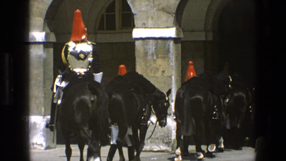 Rear view of ceremonial mounted guards and black horses moving toward shadowed stone archways in 1969 England footage.