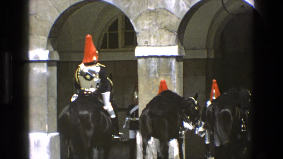 Ceremonial mounted guards on black horses cluster near a shaded stone archway in 1969 England home-movie footage.