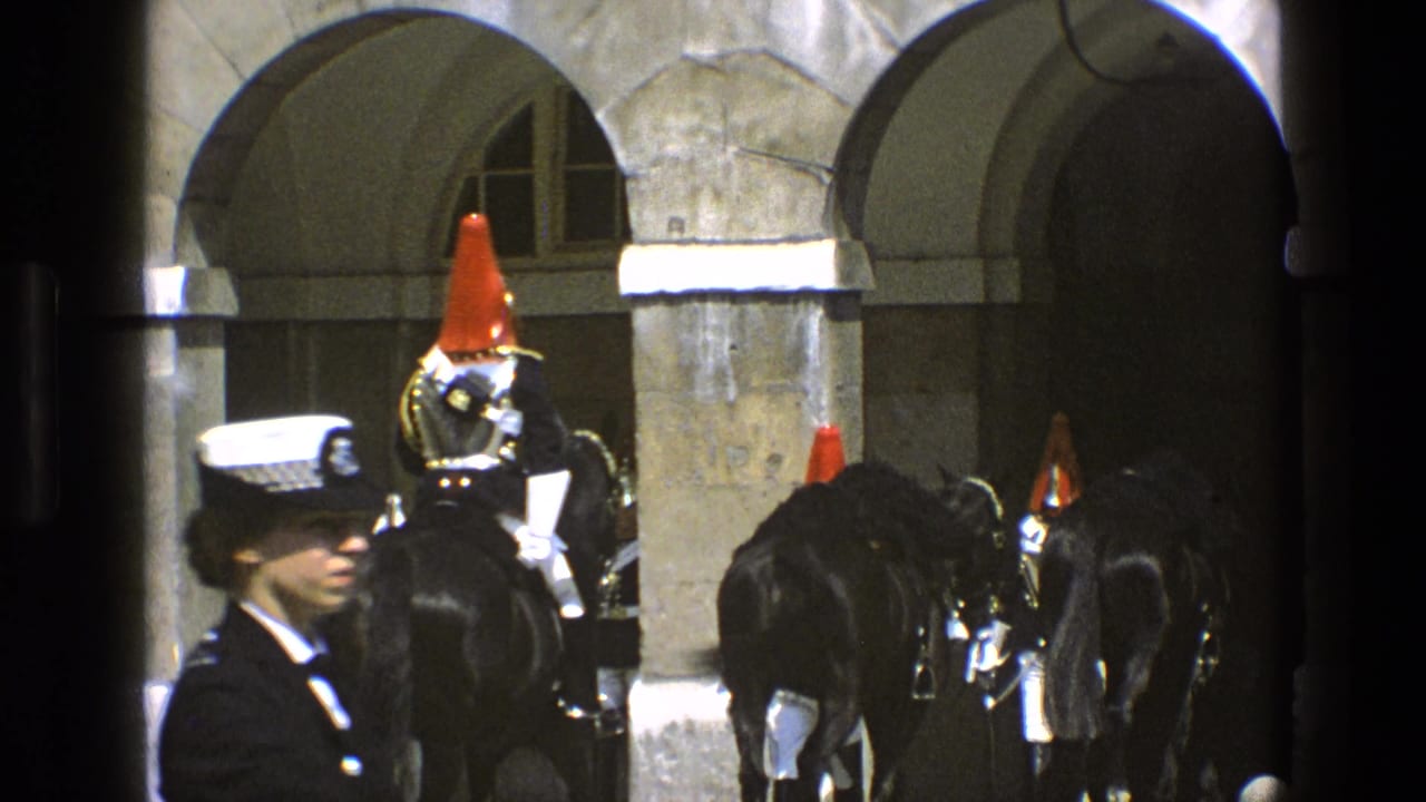 In England archival 8mm footage from 1969, mounted ceremonial guards ride black horses through stone arches while a police officer watches nearby.