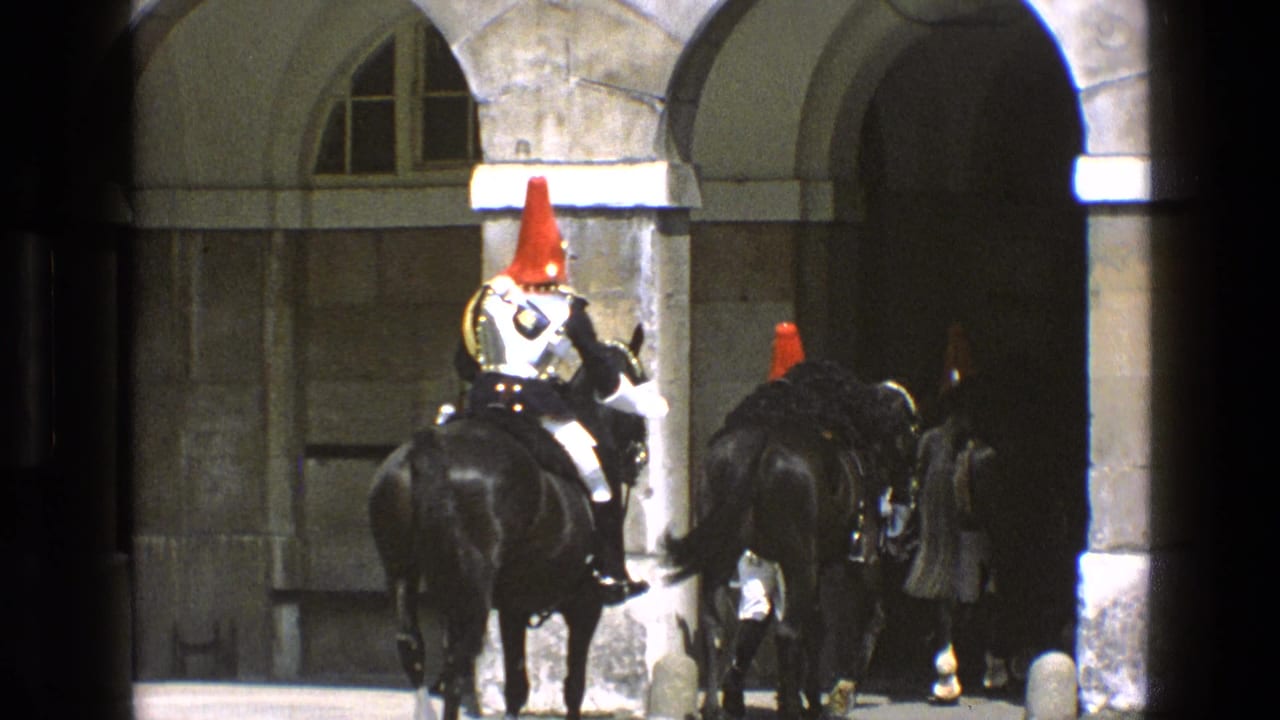 Mounted ceremonial guards with red helmet plumes and polished armor ride black horses through a stone archway in 1969 England footage.