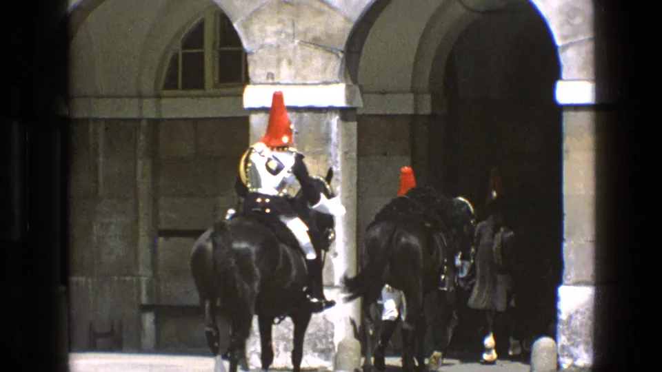Mounted ceremonial guards with red helmet plumes and polished armor ride black horses through a stone archway in 1969 England footage.