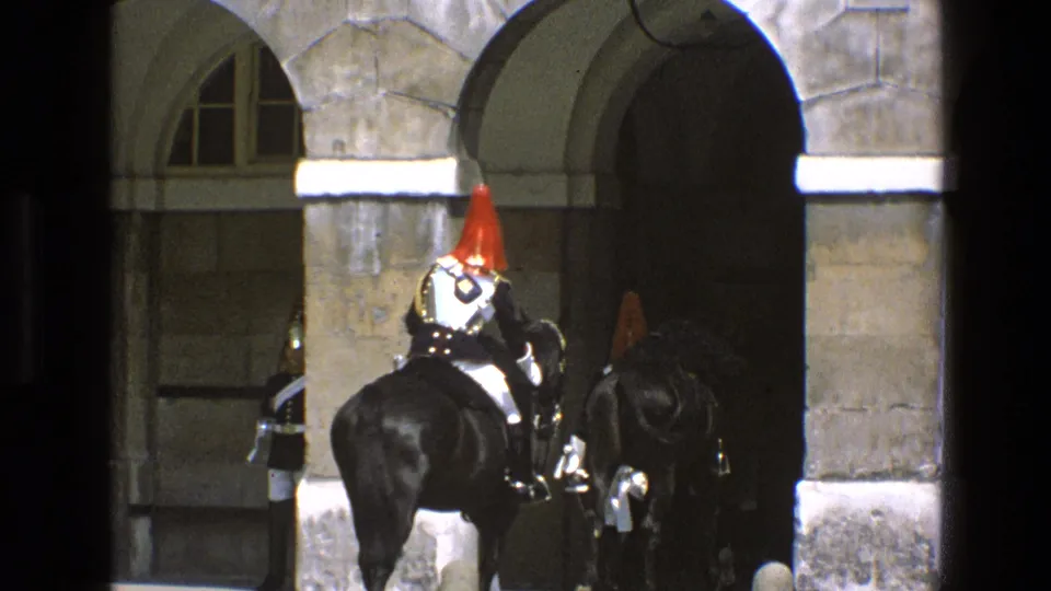 A guard in ceremonial armor and red plume rides a black horse near a stone arch, with another mounted figure partly visible beside him.