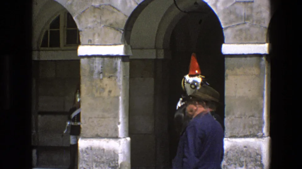 A civilian in a hat stands near the arch as the red-plumed mounted guard is partly visible in England 1969 footage.