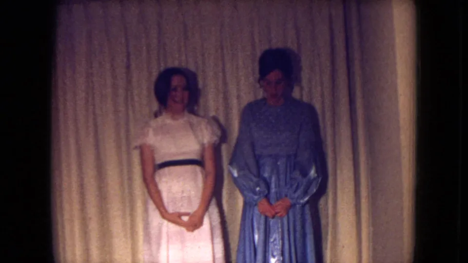 Two women pose indoors in Fort Wayne, Indiana, one in a white dress with a dark belt and one in a blue gown, in 1969 home-movie footage.