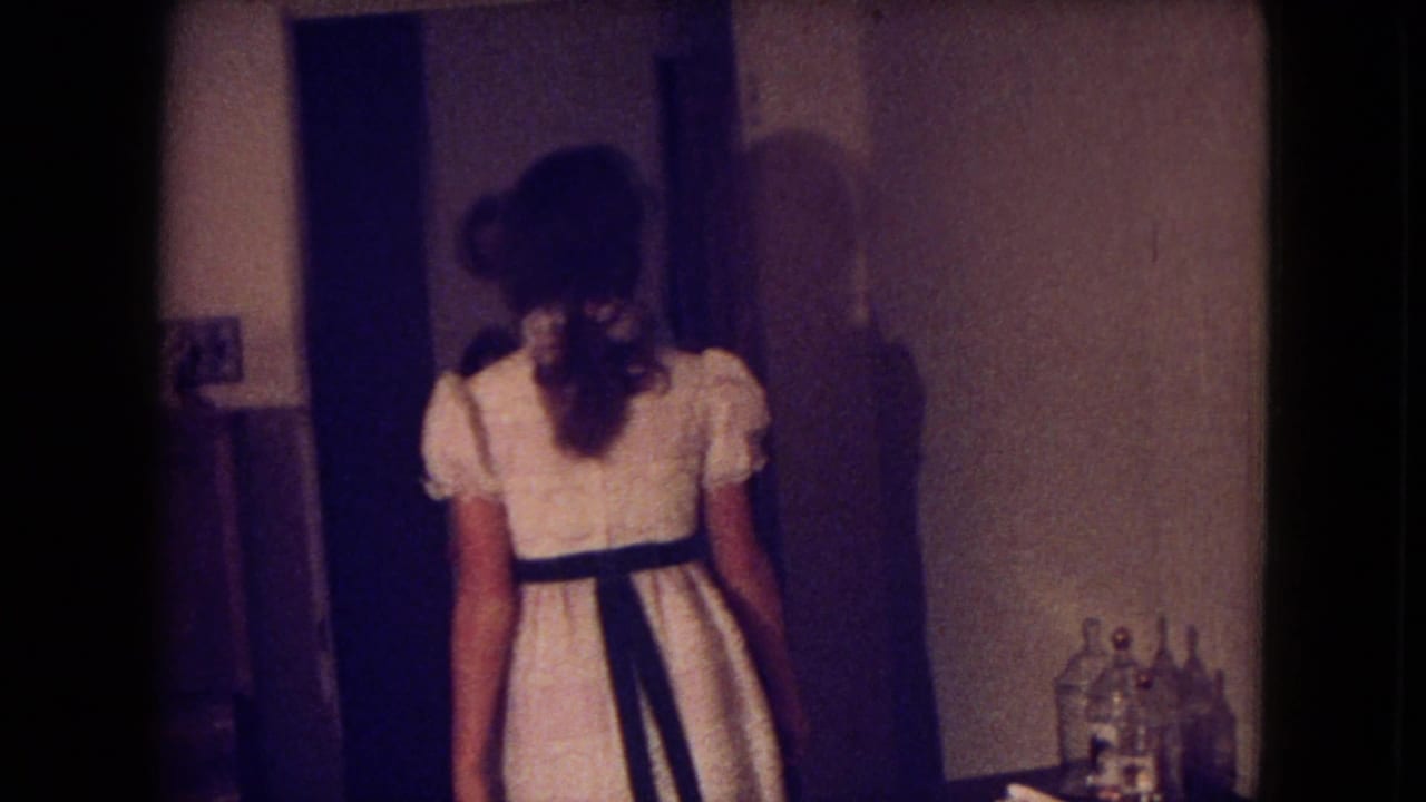 A woman in a white dress with a dark belt walks through an interior doorway in the Fort Wayne 1969 home-movie clip.