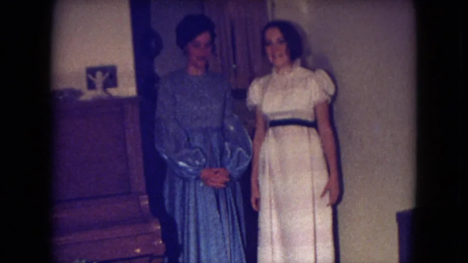 In Fort Wayne home-movie footage from 1969, two young women pose indoors near a piano, one in blue and one in white.