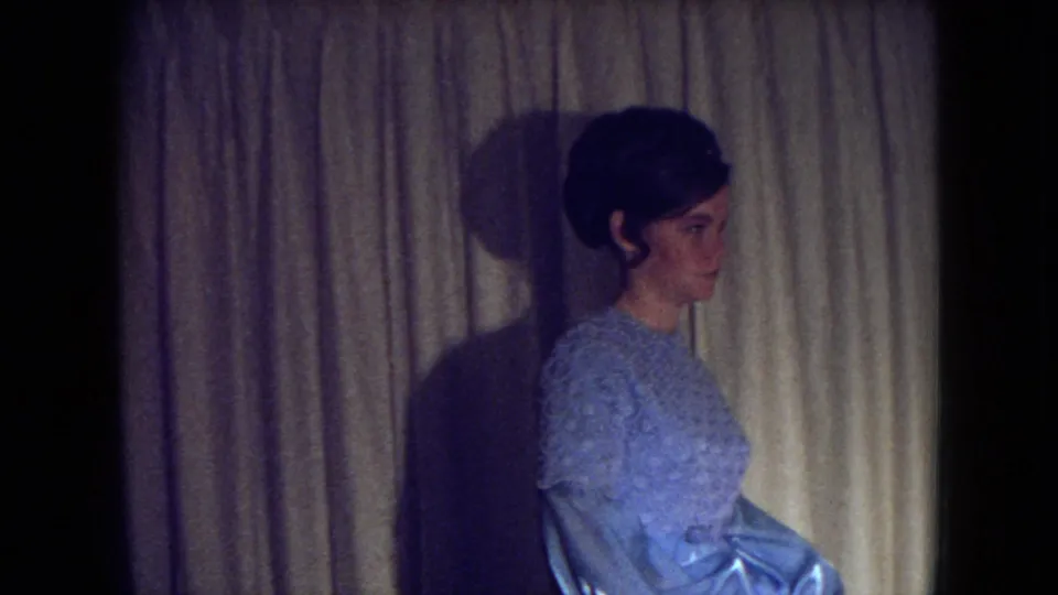 A young woman in a shimmering blue gown appears in profile against curtains in the 1969 Fort Wayne home-movie clip.