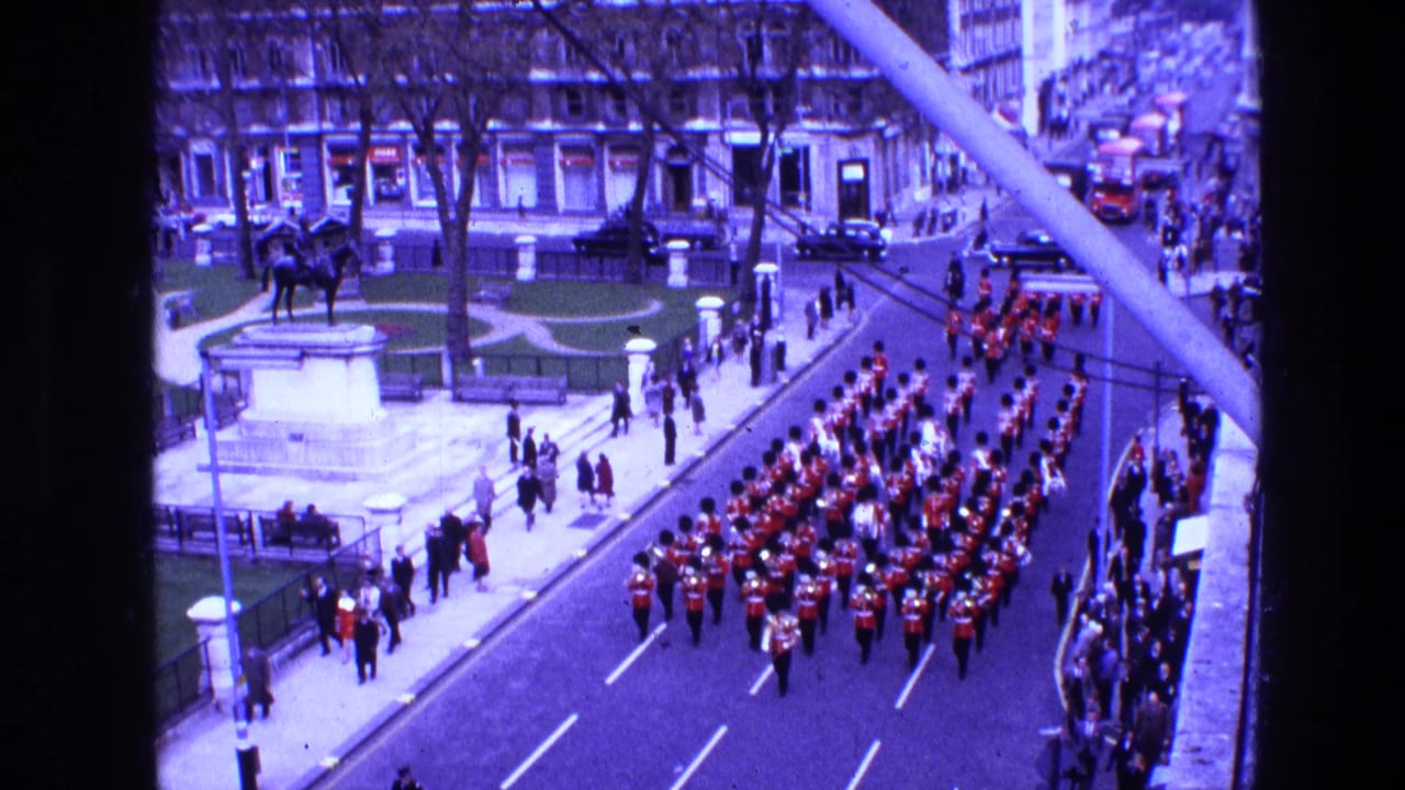 Marching band in red uniforms parades past spectators beside a small park and equestrian statue.