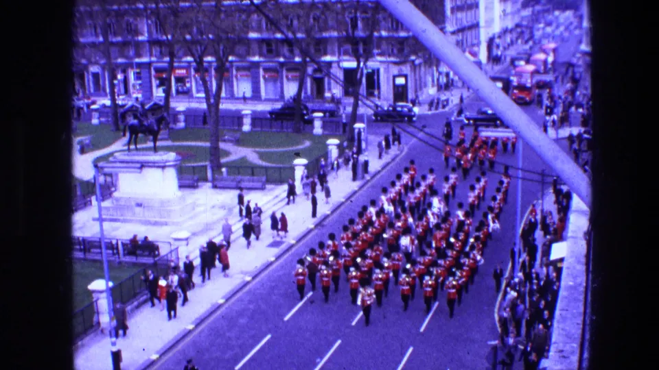 Elevated 1969 Ireland 8mm view of a red-uniformed marching band passing a crowd near a fenced green space and equestrian statue.