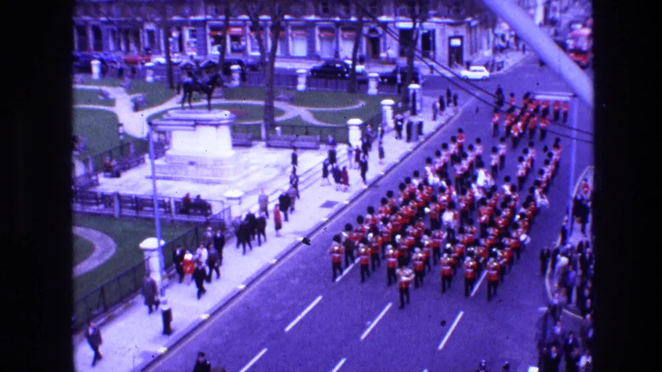 Elevated 8mm parade view in Ireland, 1969, showing a large uniformed band marching past crowds, railings, trees, and stone-fronted buildings.