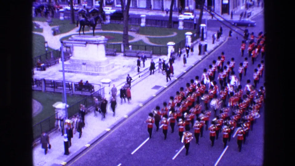 Final 1969 Ireland parade frame shows the red-uniformed band continuing down the street as pedestrians and spectators cluster near park railings and an equestrian statue.