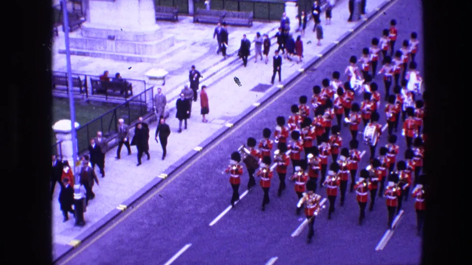 1969 Ireland home-movie frame showing a marching band in red tunics moving down a road past pedestrians and a monument base.