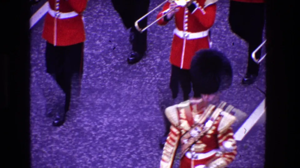 1969 8mm high-angle view of ceremonial marching musicians in red tunics and tall black hats, with a band leader partly visible below.