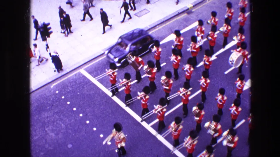 Wide 1969 8mm view of the ceremonial marching band moving along a city street as pedestrians and a dark car appear nearby.