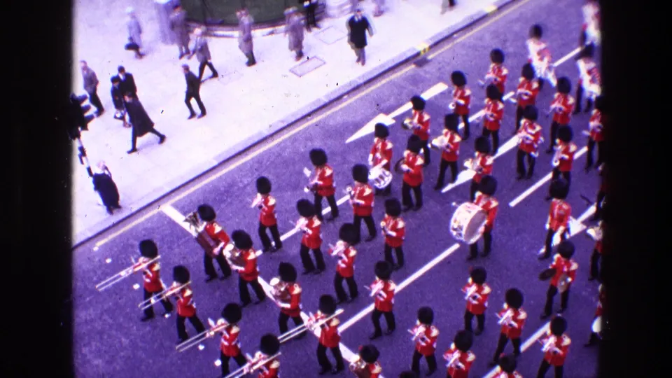 The marching band continues down the street in formation, with pedestrians on the pavement and drums and brass instruments visible.