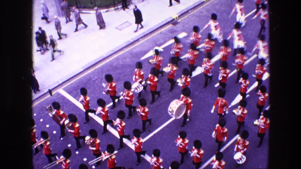 High-angle 1969 Ireland 8mm frame of a red-coated marching band with brass and drums passing pedestrians on a city street.