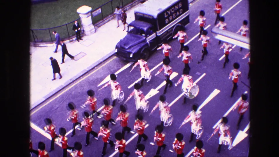 1969 Ireland parade frame with a Lyons Bread van, pedestrians, and red-uniformed band members moving through traffic lanes.