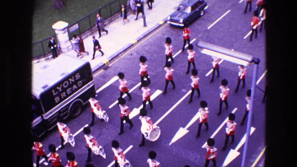 High-angle 1969 Ireland 8mm view of red-coated band members, a Lyons Bread van, pedestrians, and street traffic.